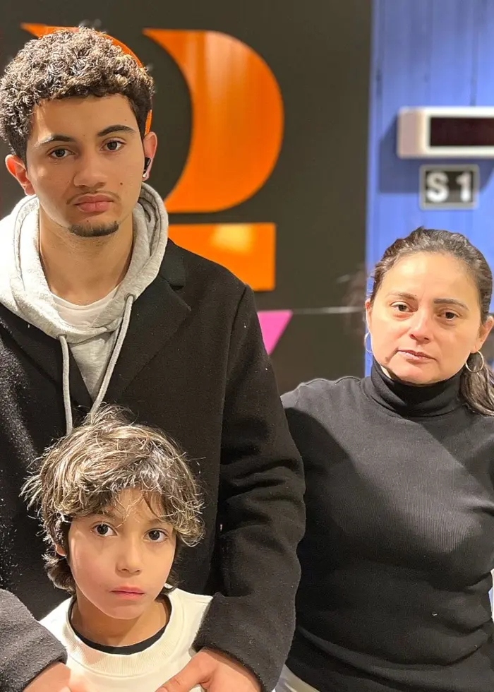 Fyra personer står tillsammans framför en färgglad vägg med text. En av dem håller ett barn i famnen. Miljön verkar vara inomhus, med en dörr och en skylt i bakgrunden.