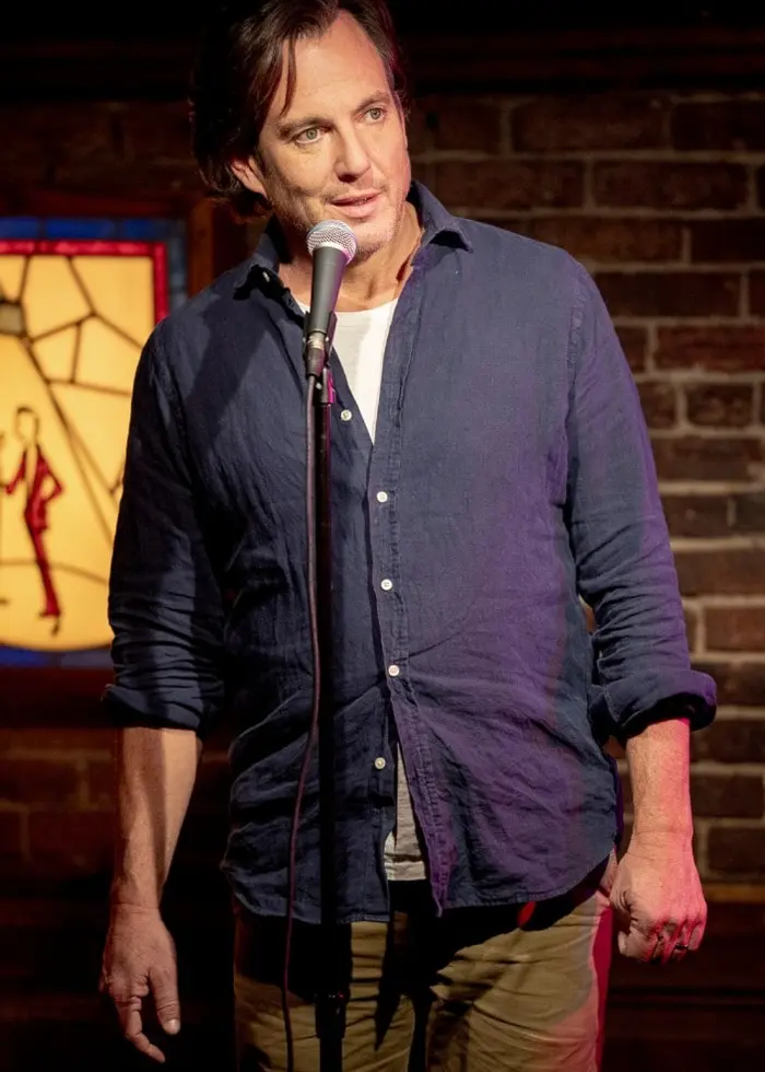 En person står på en scen framför en mikrofon i en mörk humorlokal. Bakgrunden visar en färgglad skylt med texten 'Comedy Cellar' och en tegelvägg.