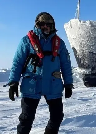 En person klädd i vinterkläder står framför ett större fartyg som är täckt av is och ligger fast i ett snötäckt landskap på Svalbard. Himlen är klarblå och ljuset skarpt.