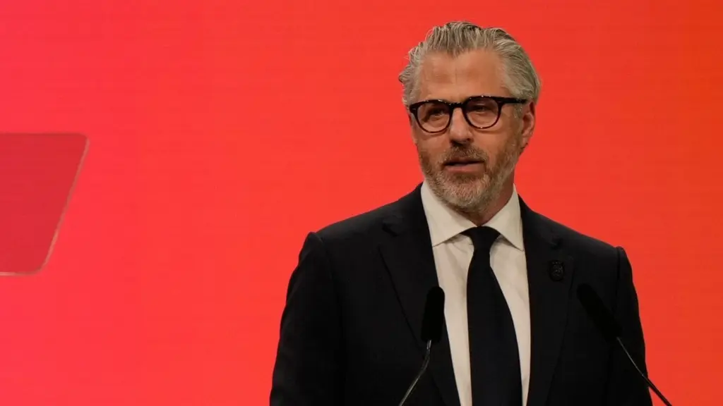 En person står vid ett podium med en bakgrund i rött och en flagga med OS-ringarna synlig. Personen håller ett tal under ett evenemang relaterat till vinter-OS 2026 i Milano, Italien.