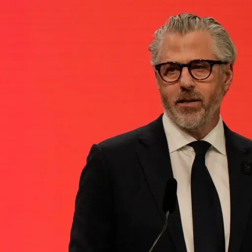 En person står vid ett podium med en bakgrund i rött och en flagga med OS-ringarna synlig. Personen håller ett tal under ett evenemang relaterat till vinter-OS 2026 i Milano, Italien.