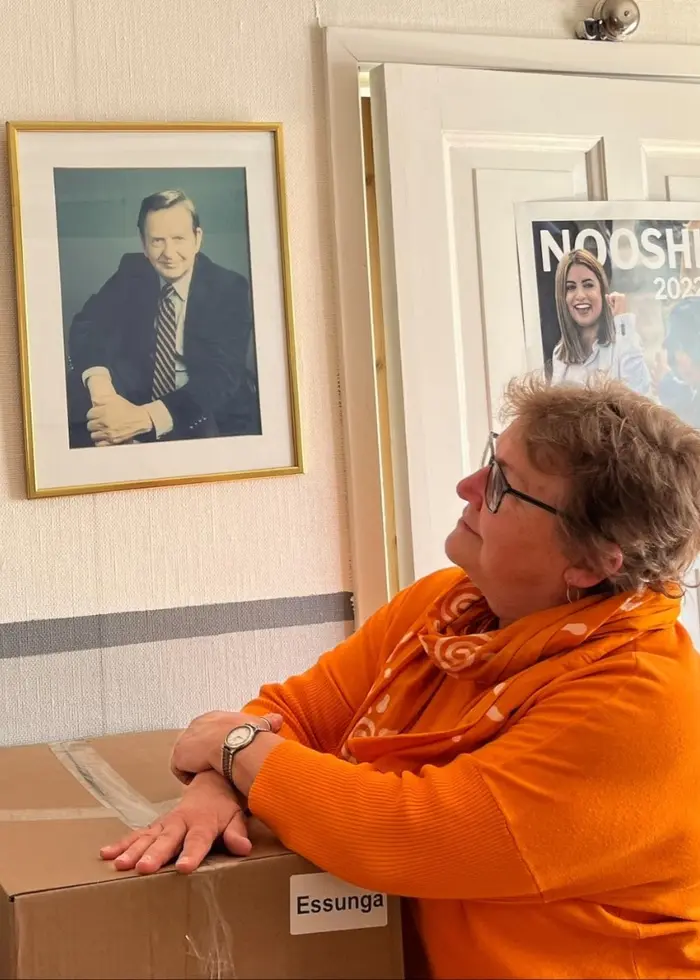 Maria Radivoj i orange tröja lutar sig mot en kartong i en S-lokal, omgiven av inramade porträtt och affischer på väggen.
