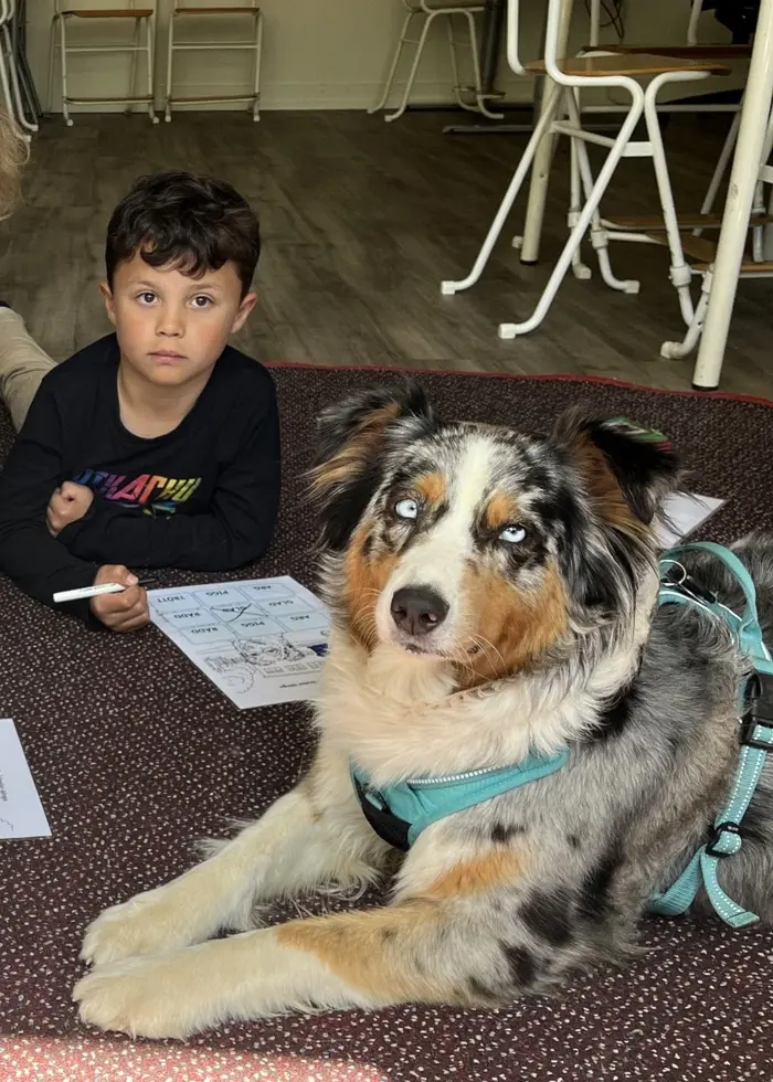 Två elever sitter på golvet bredvid en hund i ett klassrum. Deras lärare sitter bredvid.